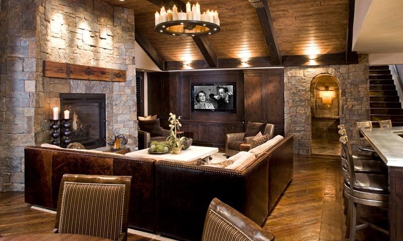 Rustic luxury media room with a leather sectional, stone fireplace, wall-mounted TV, and warm layered lighting.