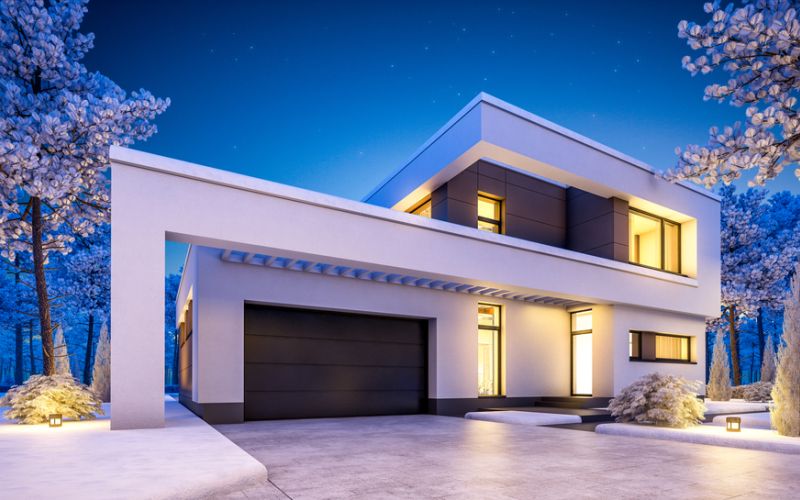 <span class="uk-h4">A modern two-story home lit at night with warm interior light, path lighting, and snow-dusted trees</span>