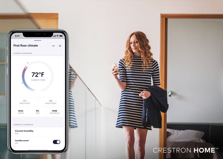 A woman in a striped dress checks her phone near a staircase, while a smartphone screen shows the Crestron Home app controlling indoor climate settings.