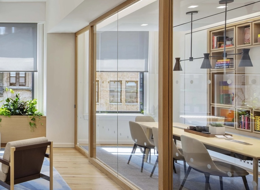 Meeting room shown through floor-to-ceiling glass sliding doors. Automated shades are partially lowered in the background.
