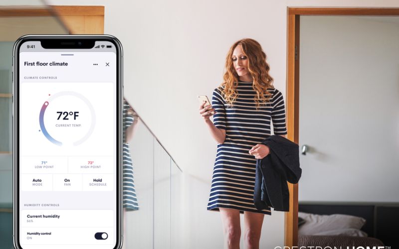 <span class="uk-h4">A woman in a striped dress checks her phone near a staircase, while a smartphone screen shows the Crestron Home app controlling indoor climate settings.</span>